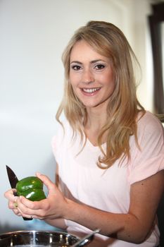 Beautiful young blond woman cutting a green bell pepper over a steel saucepan in the kitchen