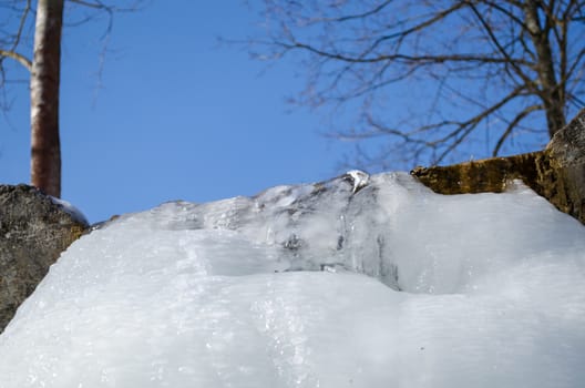 small park stream waterfall cascade covered with ice sulight and water flow under it on background of blue sky.