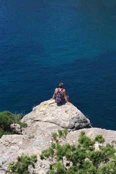 Lonely young hiker sitting on top of the rock and looking to the blue sea