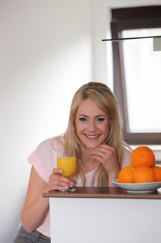 Smiling woman drinking a glass of freshly squeezed orange juice in her kitchen alongside a large bowl of fresh oranges