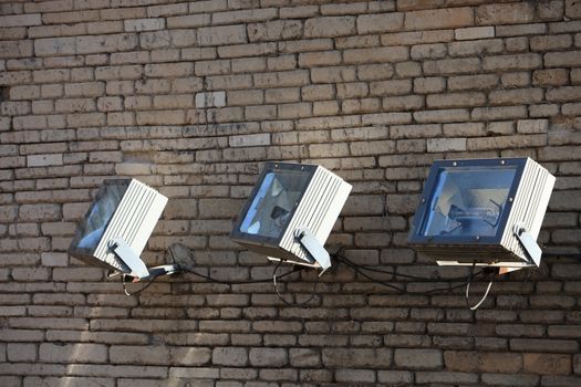 Three spotlights on an exterior brick wall mounted angled upwards to illuminated a building or landmark