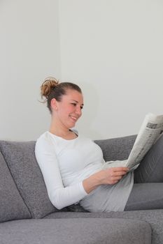 Attractive young woman enjoying her morning newspaper relaxing on the sofa in her living room with copyspace