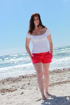 Full length portrait of a barefoot attractive woman in shorts standing on the beach in front of the blue ocean on a hot summer day