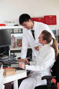 Two laboratory technicians, a young Asian man and Caucasian woman, discussing their work at a bench in a modern chemical testing laboratory
