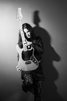 Photo of a female guitarist leaning up against a wall holding an electric guitar.
