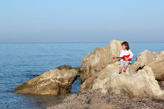 little girl sitting on a rock by the sea and playing guitar