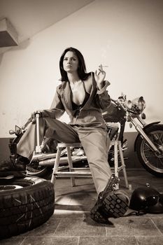 Photo of a beautiful female mechanic wearing overalls, leather bra, and sitting in front of a motorcycle smoking and holding a tool.