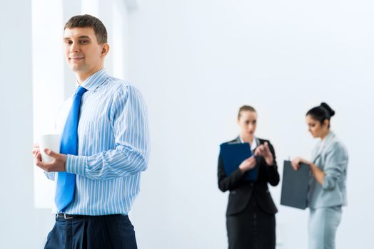 portrait of a successful businessman, holding a white mug, in the background talking colleagues