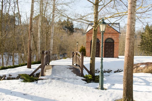 park bridge through brook stream retro lighting pole lamp and red brick house surrounded by snow in winter.