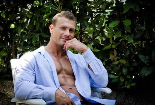 Attractive young muscle man smiling, outdoors, sitting and showing muscular pecs, abs, arms and torso