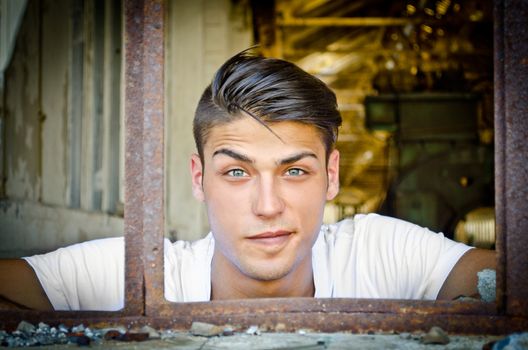 Attractive young man at rusty window with funny expression on his face, looking in camera