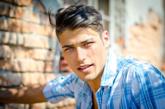 Attractive young man outdoors smiling and looking in camera, blue shirt, in front of brick wall