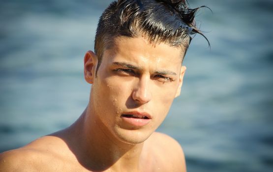 Attractive young man in the sea getting out of water with wet hair, looking in camera