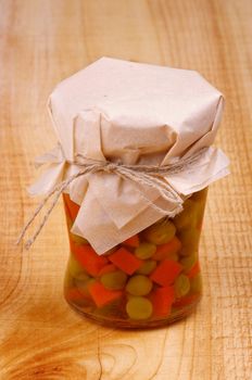 Glass Jar with Homemade Pickled Pieces of Carrot and Green Pea Covered Butter-Paper on Wooden background