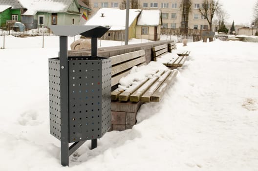 wooden park benches and waste bin covered with snow. winter town fragment and lithuania flag on house.