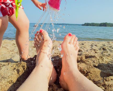 Girl washing mothers red pedicure feet with watering can at beach