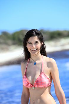 Joyful young woman at the beach standing smiling in her bikini in front of a calm ocean in the summer sun