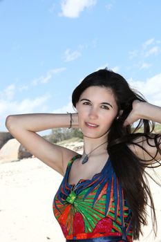 Beautiful woman on a summer vacation at the beach posing with her hands raised behind her neck in her long straight brunette hair wearing a colourful summer dress