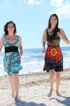 Two attractive barefoot women on the beach in their colourful summer dresses walking fast towards the camera across the sand with the sea behind them