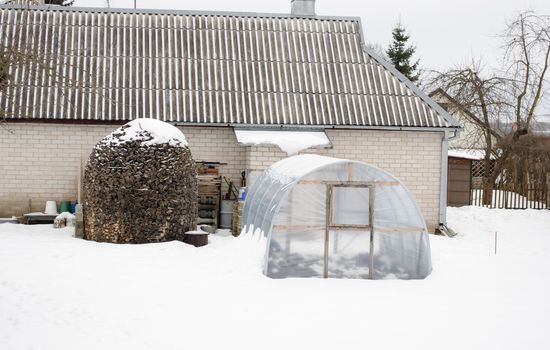 stack of firewood wood and polythene greenhouse conservatory covered with snow near white brick house in winter.