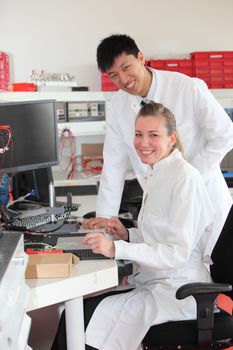Two technicians at work in a laboratory with a young Asian man standing over his attractive female colleague as they discuss a test result