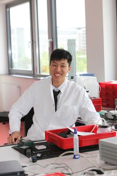 Smiling Asian man working in a laboratory sitting at his desk in a white lab coat with copyspace