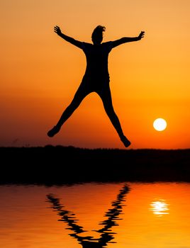 Silhouette woman jumping against orange sunset with reflection in water