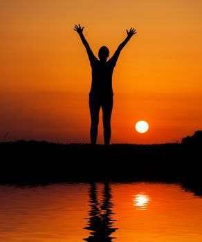 Silhouette woman jumping against orange sunset with reflection in water