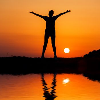 Silhouette woman jumping against orange sunset with reflection in water