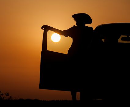 Silhouette woman with hat standing near car, against orange sunset