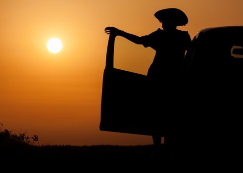 Silhouette woman with hat standing near car, against orange sunset