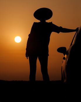 Silhouette woman with hat standing near car, against orange sunset