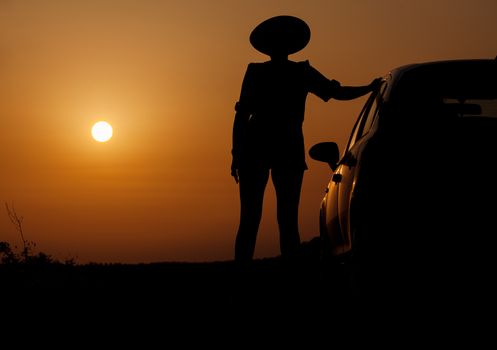 Silhouette woman with hat standing near car, against orange sunset