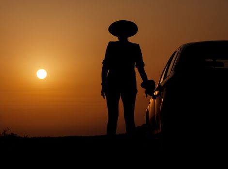 Silhouette woman with hat standing near car, against orange sunset