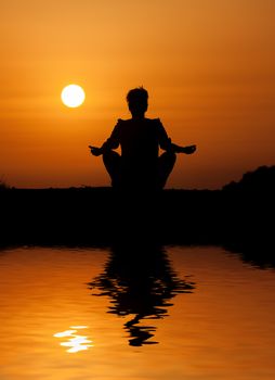 Silhouette woman sitting and relaxing against orange sunset with reflection in water