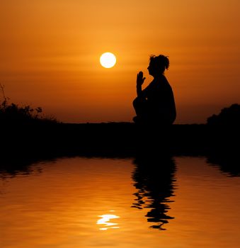 Silhouette woman sitting and relaxing against orange sunset with reflection in water