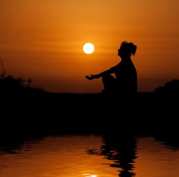 Silhouette woman sitting and relaxing against orange sunset with reflection in water