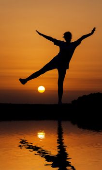 Silhouette woman jumping against orange sunset with reflection in water