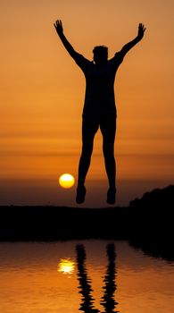 Silhouette woman jumping against orange sunset with reflection in water