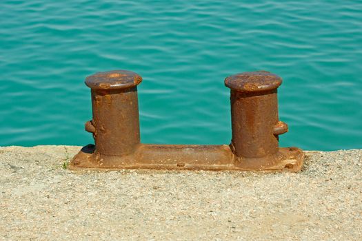 Steel rusty dual mooring bitt on a fragment of old concrete pier on the background turquoise sea water in calm