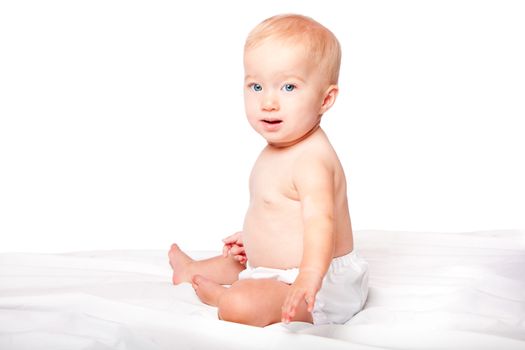 Cute blond baby with blue yes sitting on soft bed wearing diapers, on white.