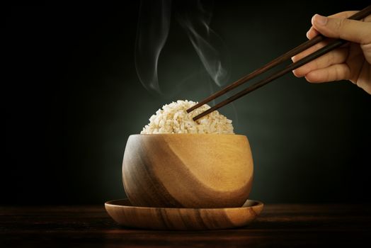 Cooked organic basmati brown rice in wooden bowl with human hand chopsticks and hot steam smoke on dining table. Low light setting.