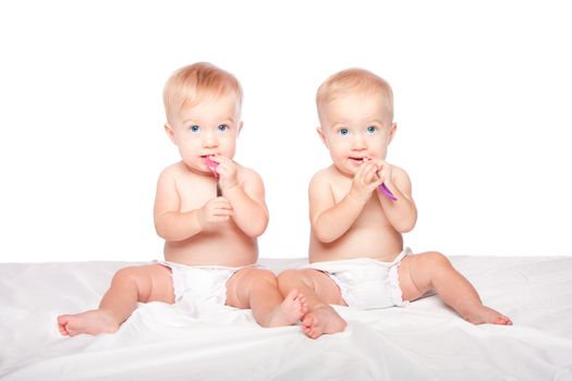 Two cute adorable infant babies with blue eyes sitting and spoons eating playing, on white.