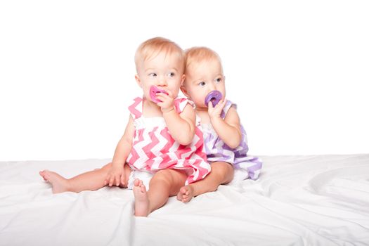 Two cute adorable identical twin girls sitting together with pacifiers, on white.