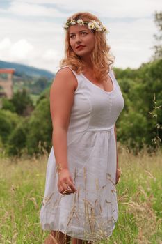 Young blond plus-size model with flower wreath in her hair and white dress standing in tall grass