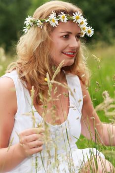 Young blond plus-size model in the white linen dress and flowers wreath sits laughing in a meadow in the high grass