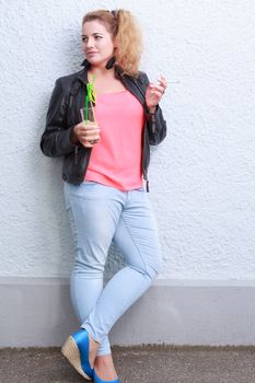 Attractive woman in jeans and a leather jacket standing drinking and smoking leaning up against a wall of a building