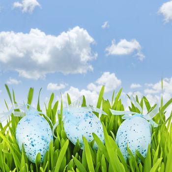 Three blue speckled easter eggs with ribbons in green grass on sky background