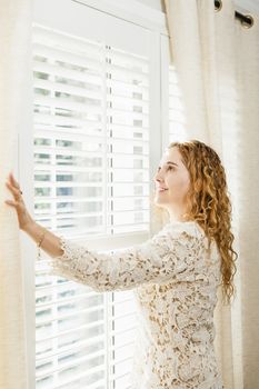 Happy woman looking out big bright window with curtains and blinds