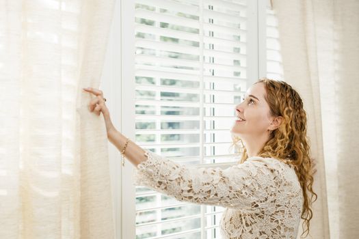 Happy woman looking out big bright window with curtains and blinds